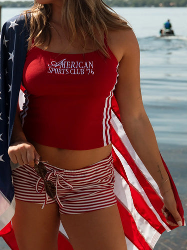 Person wearing a red sports top and striped shorts with an American flag, standing by a water body.