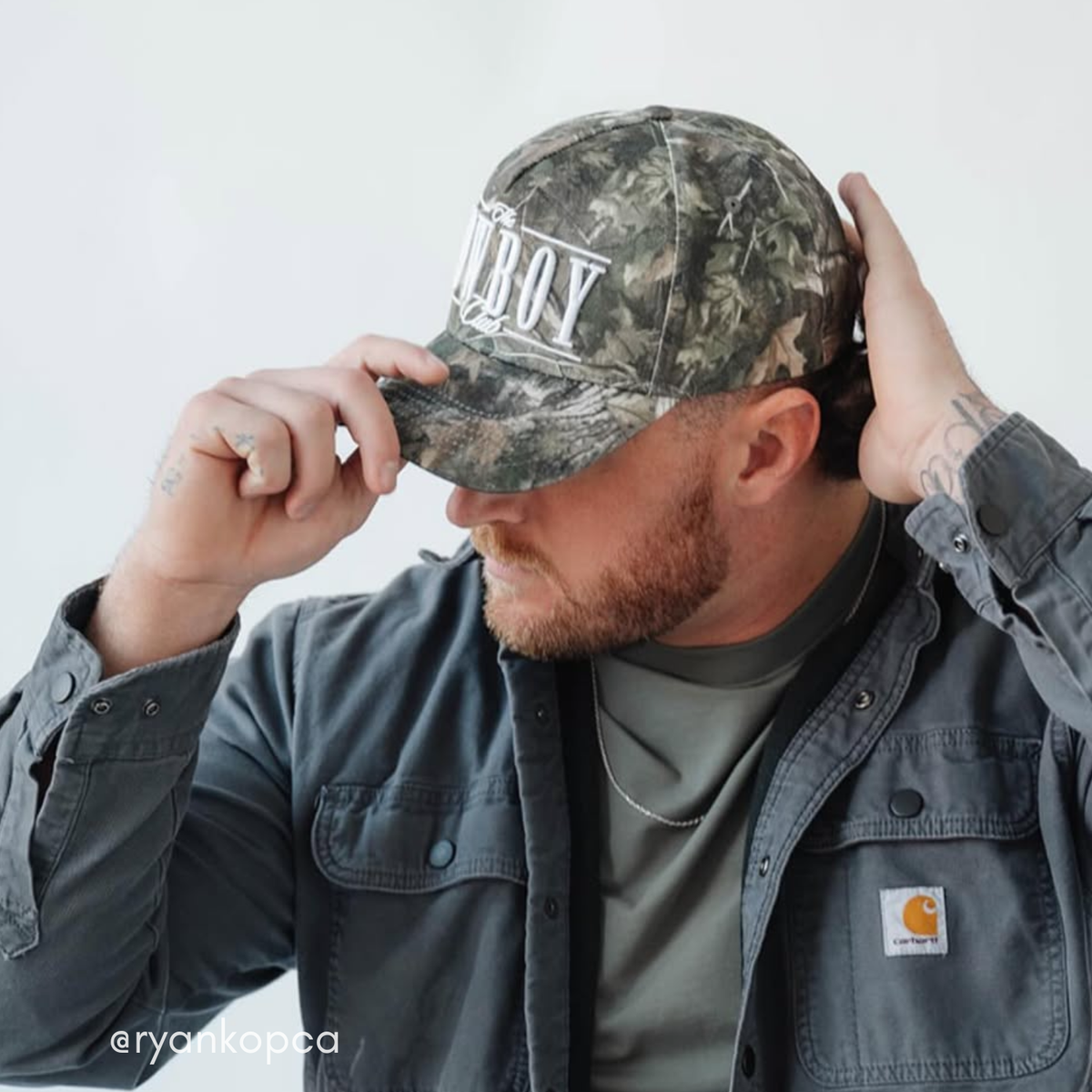 Man wearing a camouflage cap with 'COWBOY Club' printed on it, adjusting the cap against a white background.