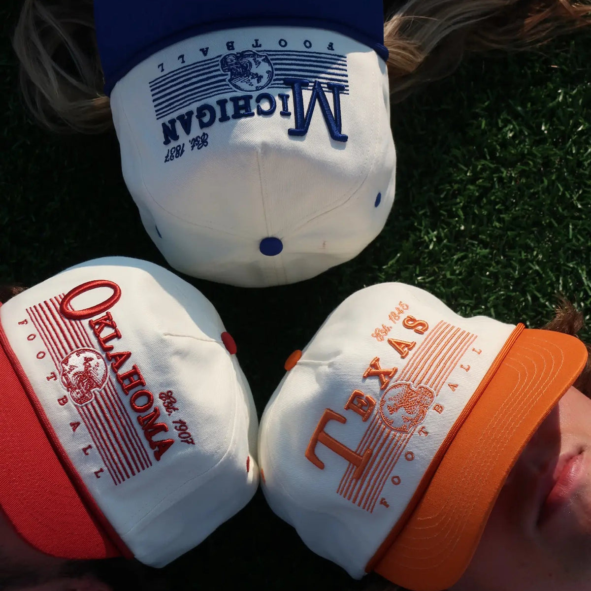 Three vintage-style baseball caps are arranged in a triangle on green grass, with parts of three people's heads visible above and below them. The top cap is white with a blue brim and blue text reading 'MICHIGAN'; the left cap is white with a red brim and red text reading 'OKLAHOMA'; the right cap is white with an orange brim and orange text reading 'TEXAS'.