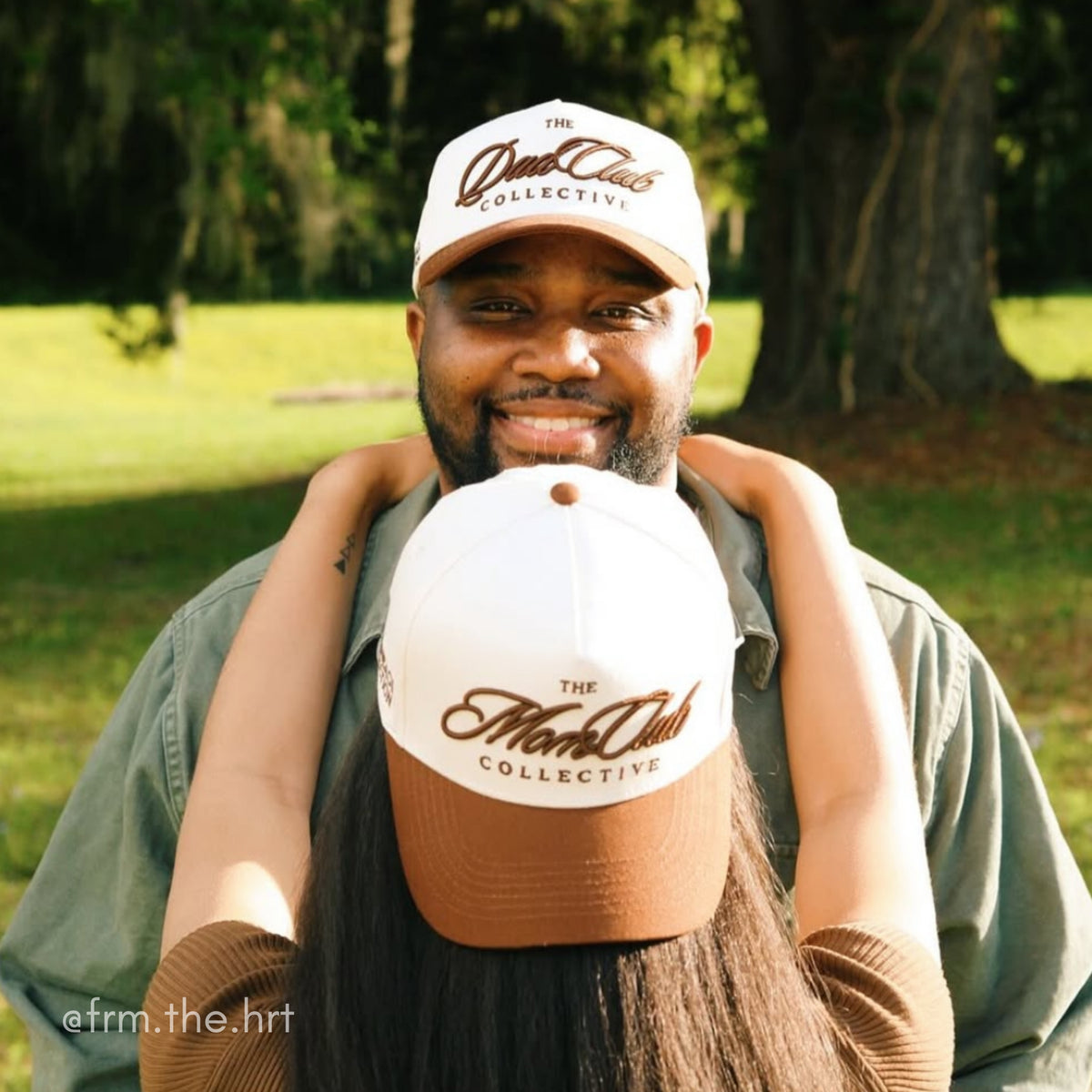 Two people wearing 'The Mom Club Collective' and 'The Dad Club Collective' hats in a park setting.
