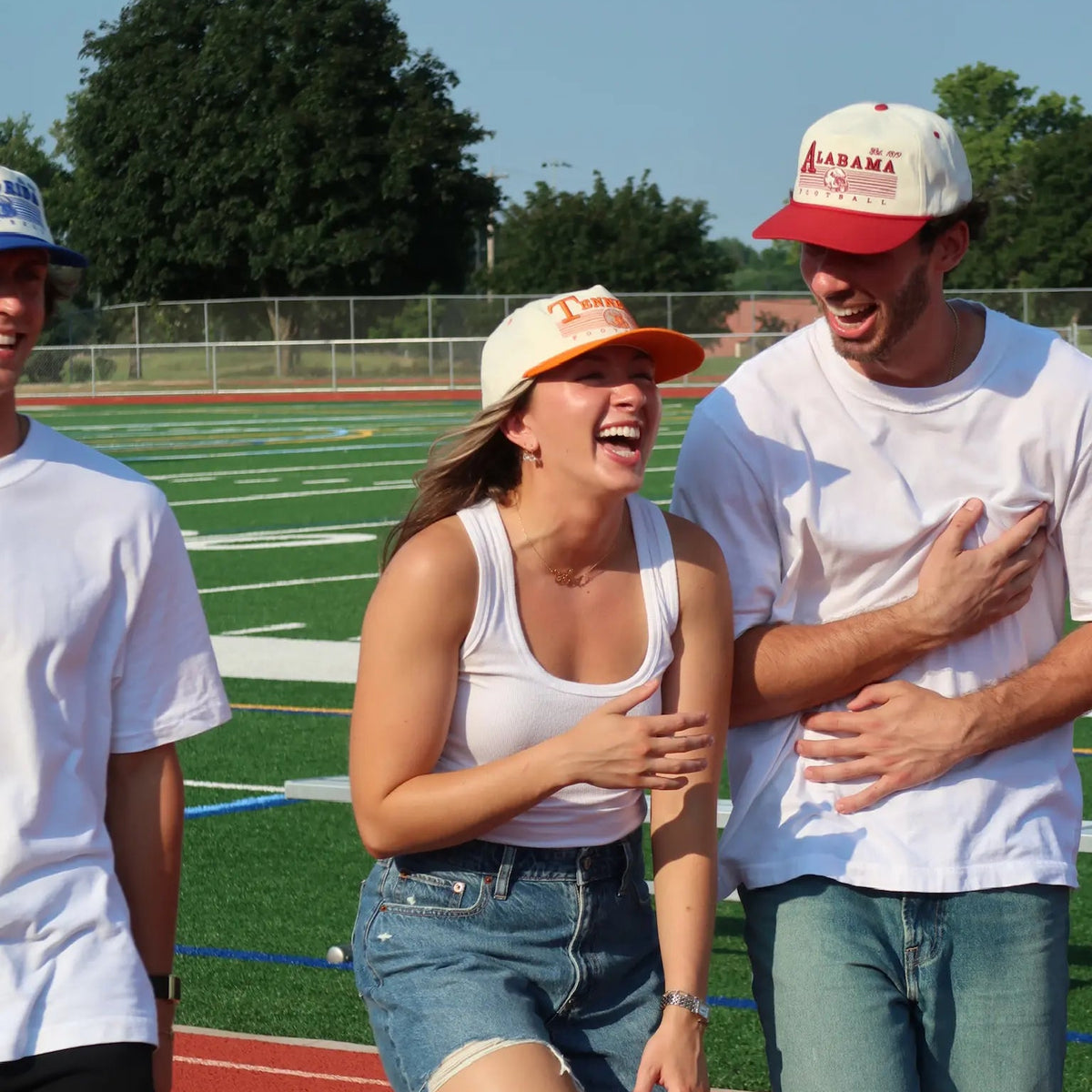 Three young adults laughing on a school track and field stadium, wearing matching white t-shirts and casual summer attire. The central woman wears denim shorts and a white tank top, while the man on the right wears a red and white Alabama baseball cap and blue jeans.